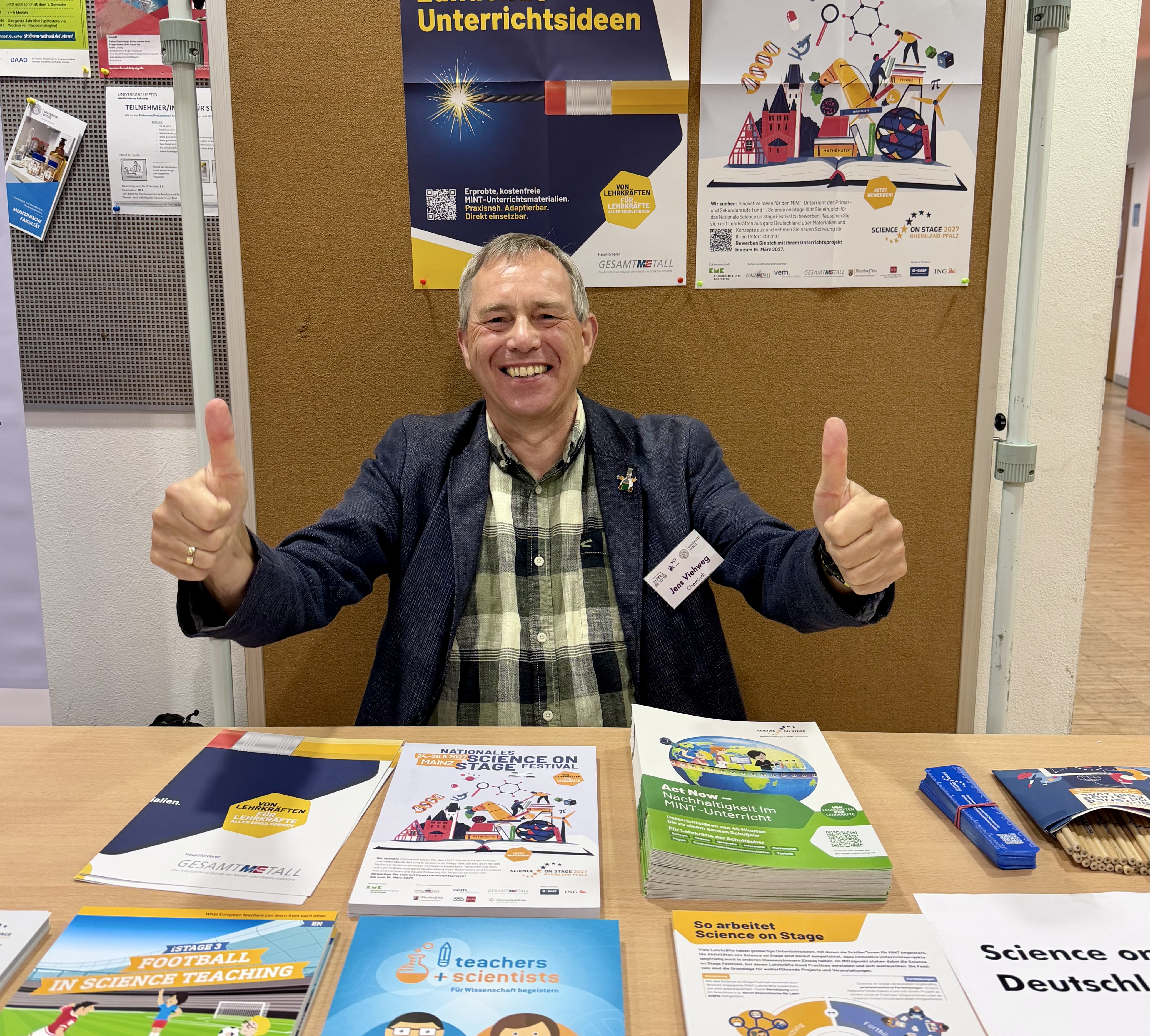 Ein Mann sitzt hinter einem Stand und zeigt mit beiden Daumen nach oben.