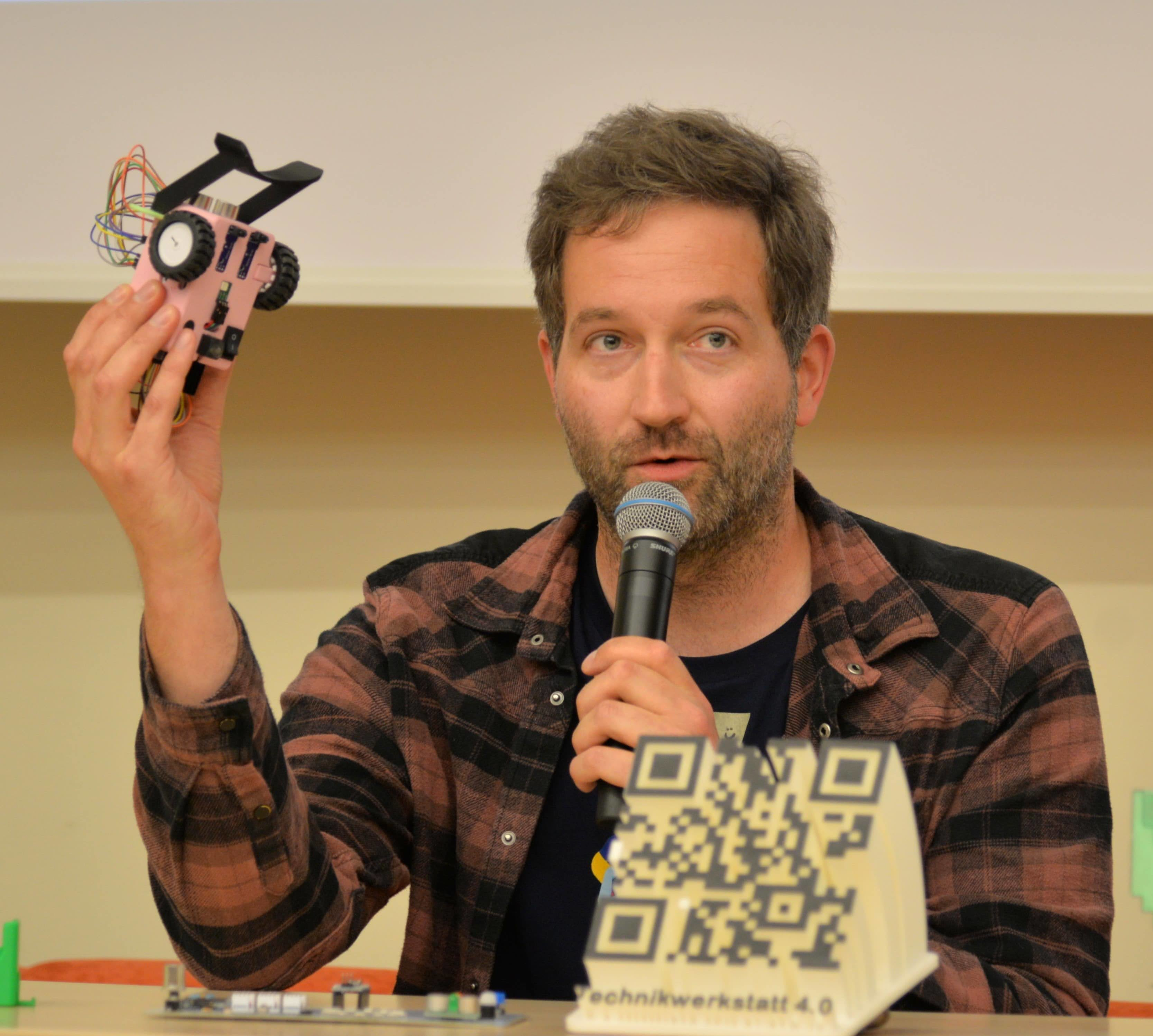 Ein Mann hält in einer Hand einen kleinen Roboter, in der anderen Hand ein Mikrofon.