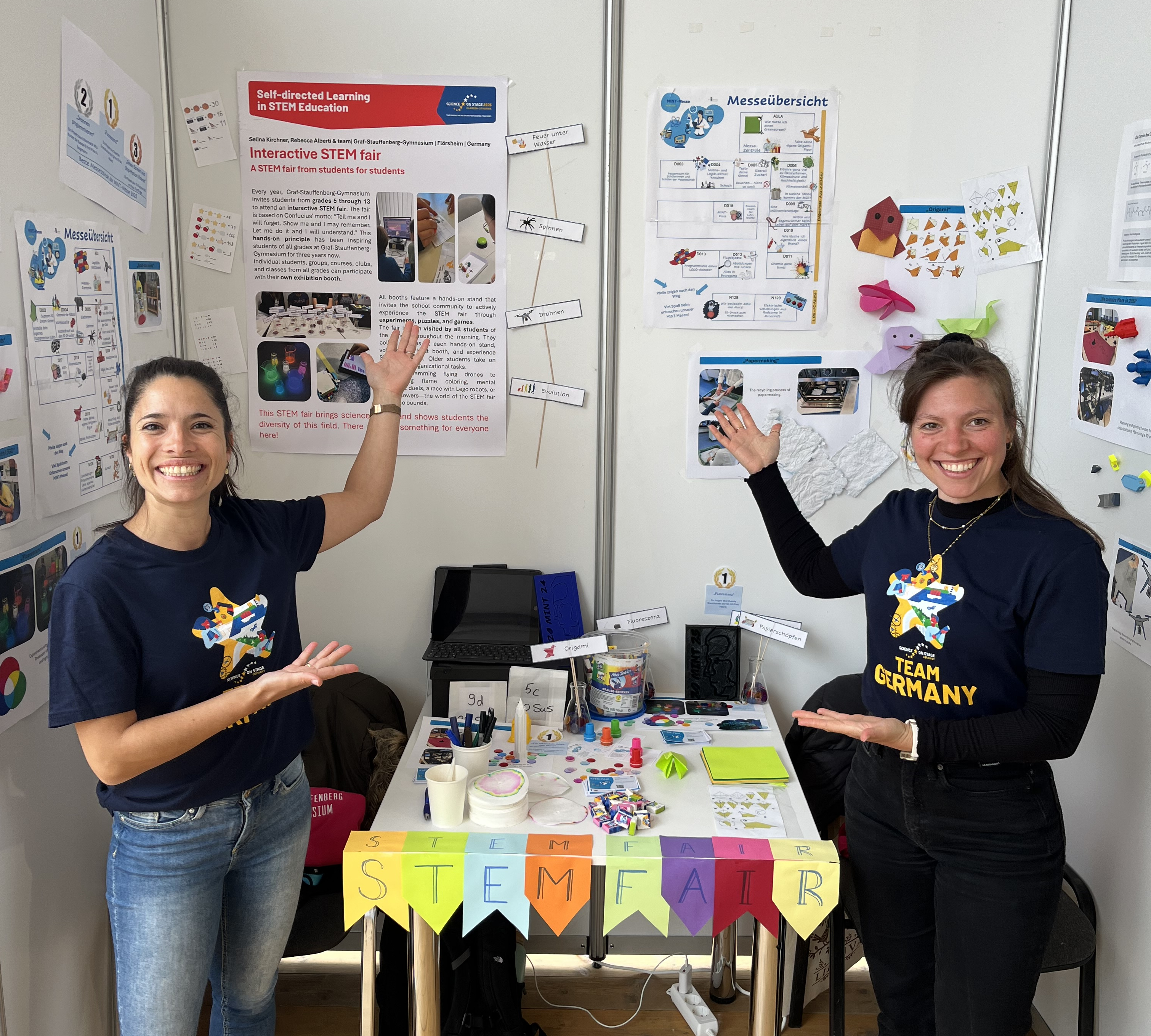 Zwei Frauen stehen an einem Stand mit Postern und einem Auslagentisch.