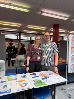 Eine Frau und ein Mann stehen an einem Stand. Auf dem Tisch liegen verschiedene Flyer und Broschüren.