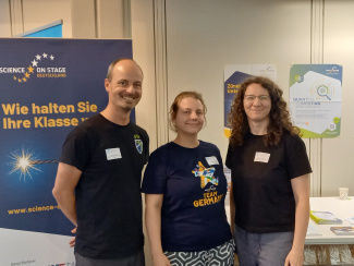 Ein Mann und zwei Frauen stehen vor einem Aufsteller und Postern.