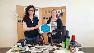 Zwei Frauen stehen an einem Stand und halten eine Plakette in der Hand