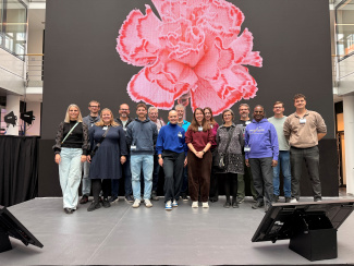 Gruppenfoto mit 15 Personen, im Hintergrund ist eine große rosa Blume auf einer schwarzen Leinwand