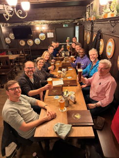 Gruppenfoto mit 13 Personen an einem langen Tisch in einer Gaststätte. Die Personen halten ihre Getränke hoch. Die Wände der Gaststätte sind mit dunklem Holz vertäfelt.
