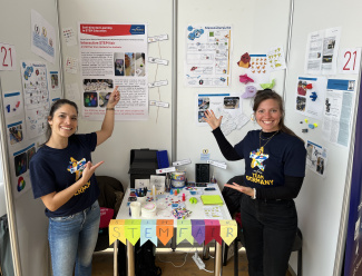 Zwei Frauen stehen an einem Stand mit Postern und einem Auslagentisch.