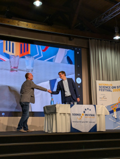 Zwei Personen schütteln sich auf einer Bühne mit Präsentationstisch die Hand