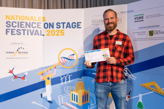 Ein Mann mit Gewinnerurkunde vor der Sponsorenwand des Science on Stage Festivals
