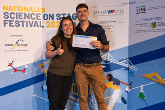 Eine Frau und ein Mann mit Gewinnerurkunde vor der Sponsorenwand des Science on Stage Festivals