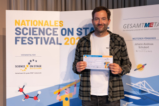EEin Mann mit Gewinnerurkunde vor der Sponsorenwand des Science on Stage Festivals
