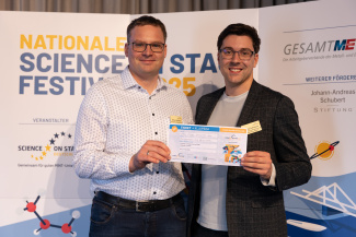 Zwei Männer mit Gewinnerurkunde vor der Sponsorenwand des Science on Stage Festivals