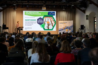 Ein voll besetzter Veranstaltungssaal mit einer Präsentation auf Großleinwand