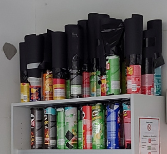 viele bunte Chipsdosen stehen in einem Regal, teilweise ist schwarze Pappe an sie angeklebt