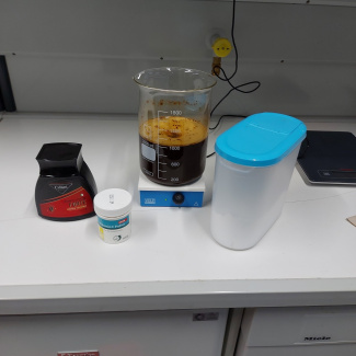 ein Becherglas mit einer dunklen Flüssigkeit, eine Dose Kaffeepulver, eine Dose Vitamin-C-Pulver, ein durchsichtiger Behälter