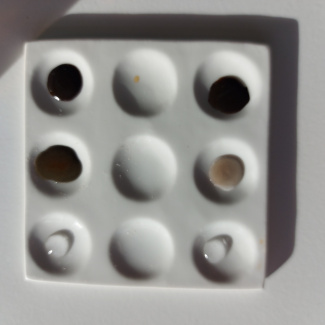 ein weiße Tüpfelplatte mit neun Vertiefungen: drei mit einer dunklen, eine mit einer hellen, zwei mit einer durchsichtigen Flüssigkeit