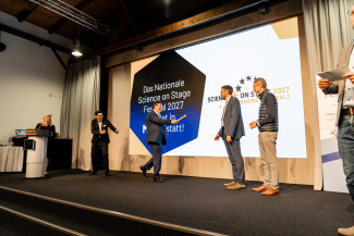 Mehrere Personen auf einer Bühne bei einer symbolischen Staffelstabübergabe