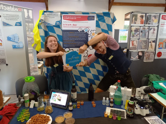 Zwei Personen in bayerischer Tracht zerren an einem Ausstellungsstand spielerisch an einer Plakette