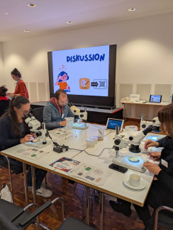 Blick auf einen Seminarraum und mehrere Personen an Mikroskopen vor einem Bildschirm mit dem Schriftzug "Discussion"